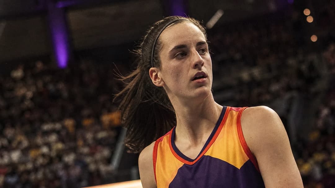 A dynamic shot of Caitlin Clark walking towards the camera in a basketball kit with fans blurred in the background.