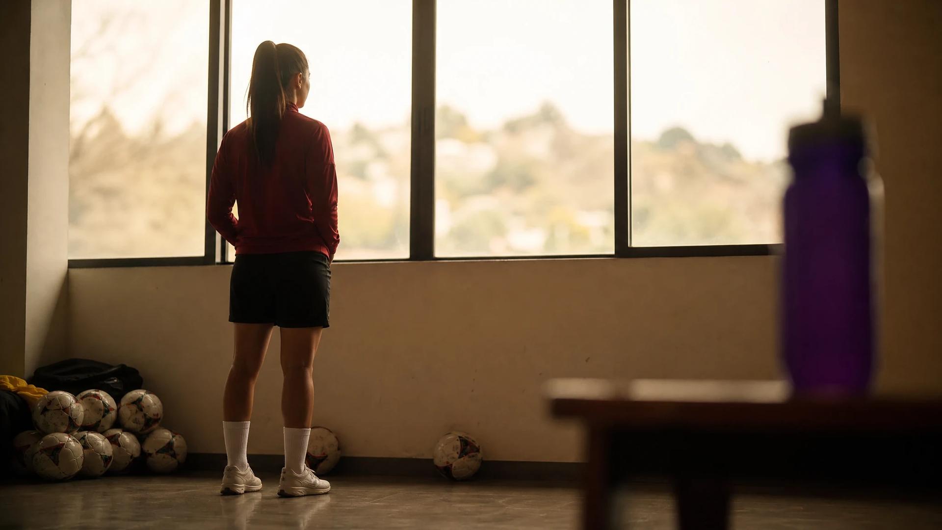 A young female footballer from Athletic stands in a training facility, looking out of a window with a violet water bottle nearby.