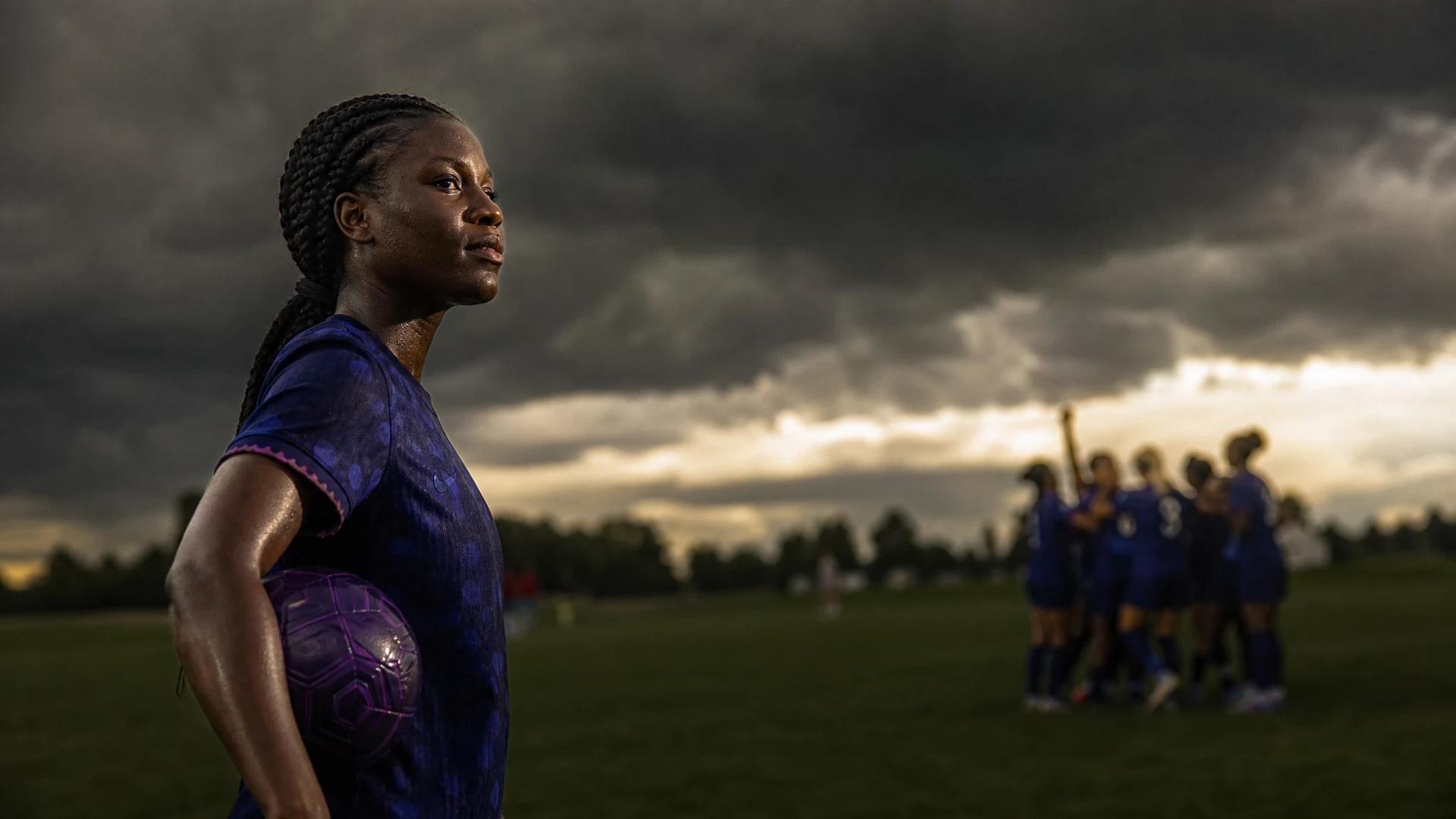 Evelyn Ijeh holding a violet soccer ball, contemplating off the pitch while teammates celebrate in the background.