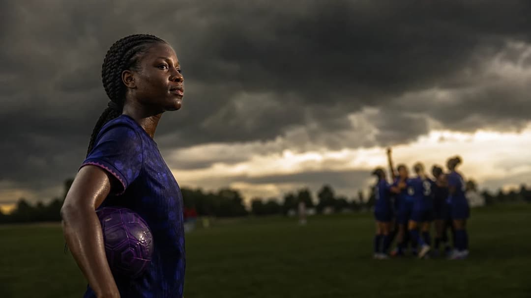 Evelyn Ijeh holding a violet soccer ball, contemplating off the pitch while teammates celebrate in the background.