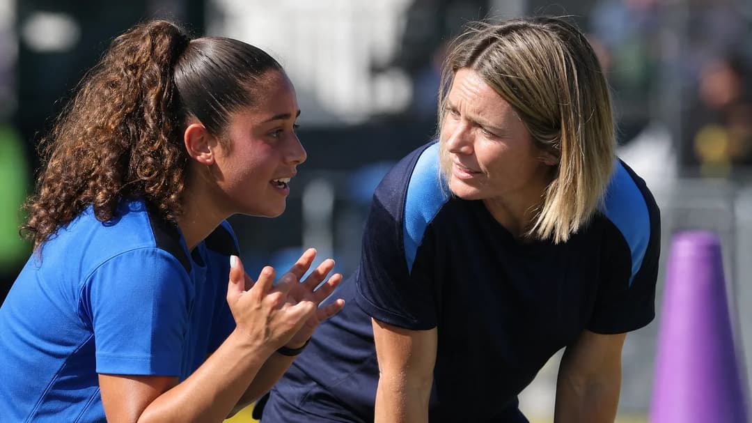 A close-up of Guilia Dragoni speaking with her coach Sonia Bompastor in bright sunlight.
