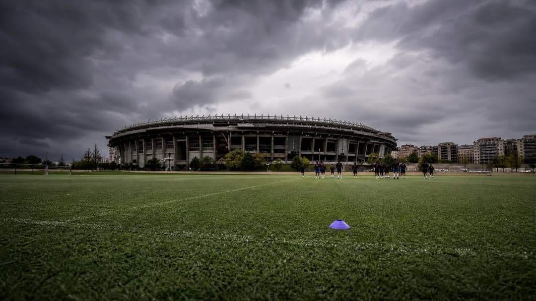 A wide shot of FC Barcelona's Camp Nou under a stormy sky, with players training in the foreground.