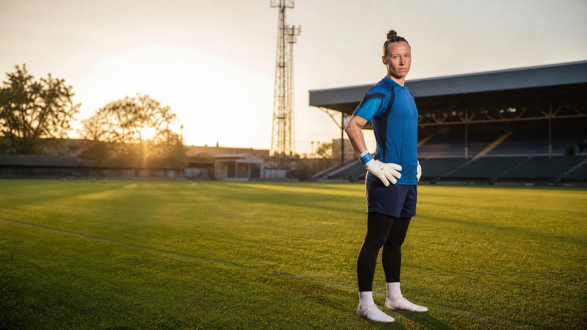 A female football goalkeeper stands on a training pitch during golden hour with a stadium in the background.