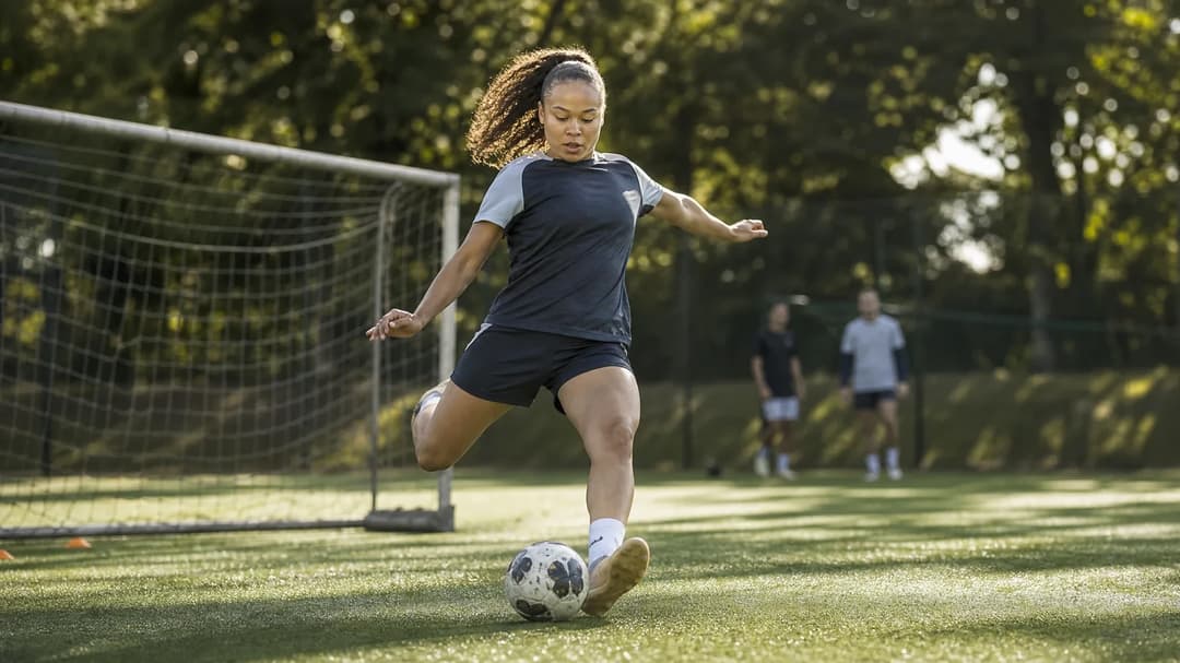 A female football player practicing a kick at a training facility, surrounded by greenery and warm sunlight.