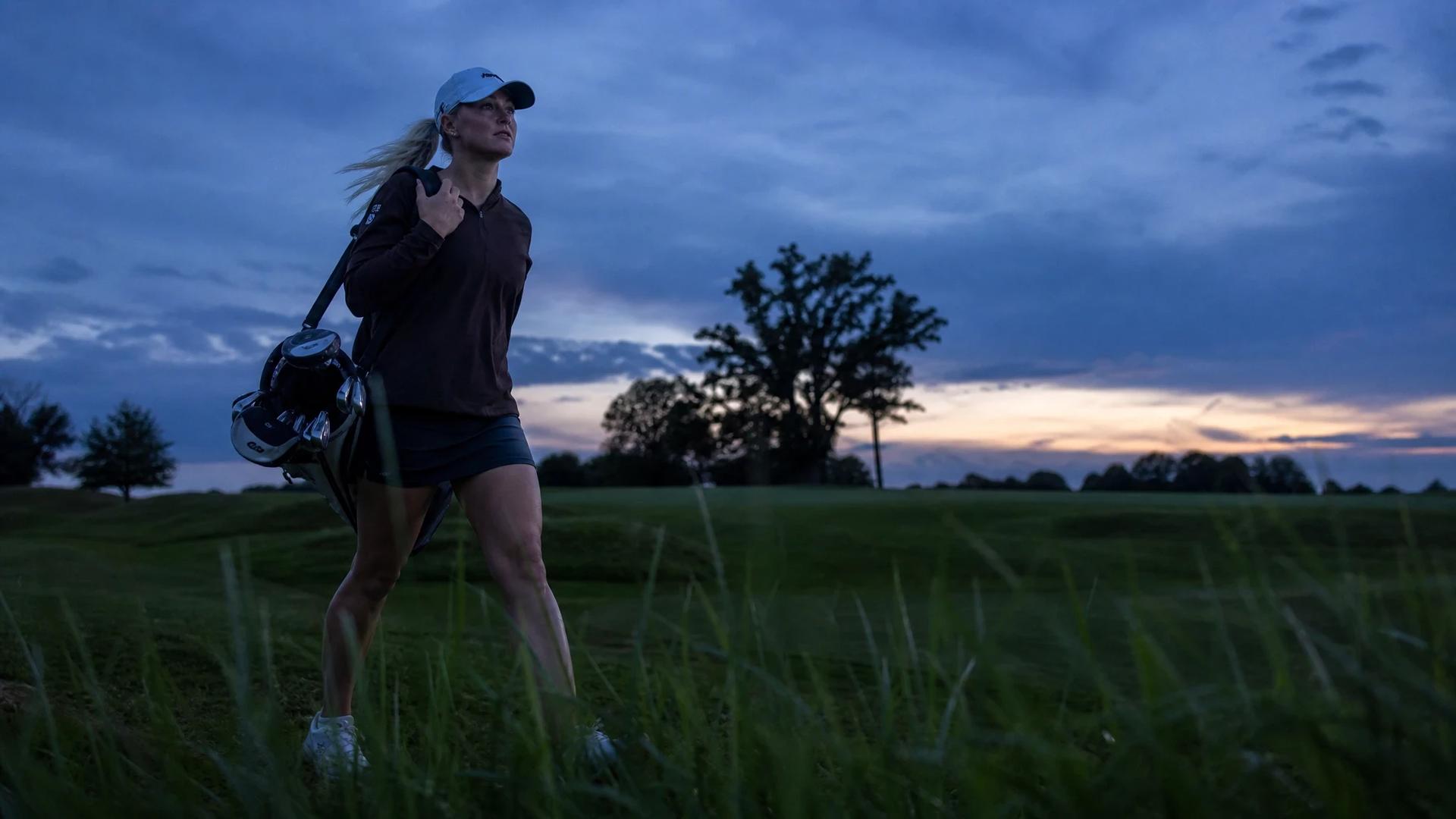 Charley Hull walking through a golf course at dusk with a golf bag over her shoulder.