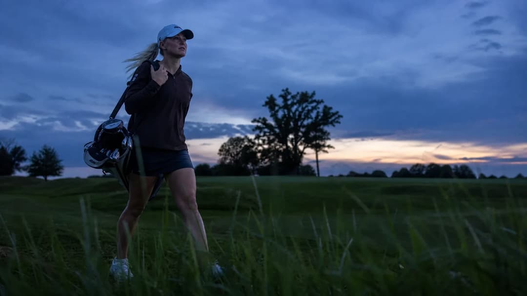 Charley Hull walking through a golf course at dusk with a golf bag over her shoulder.