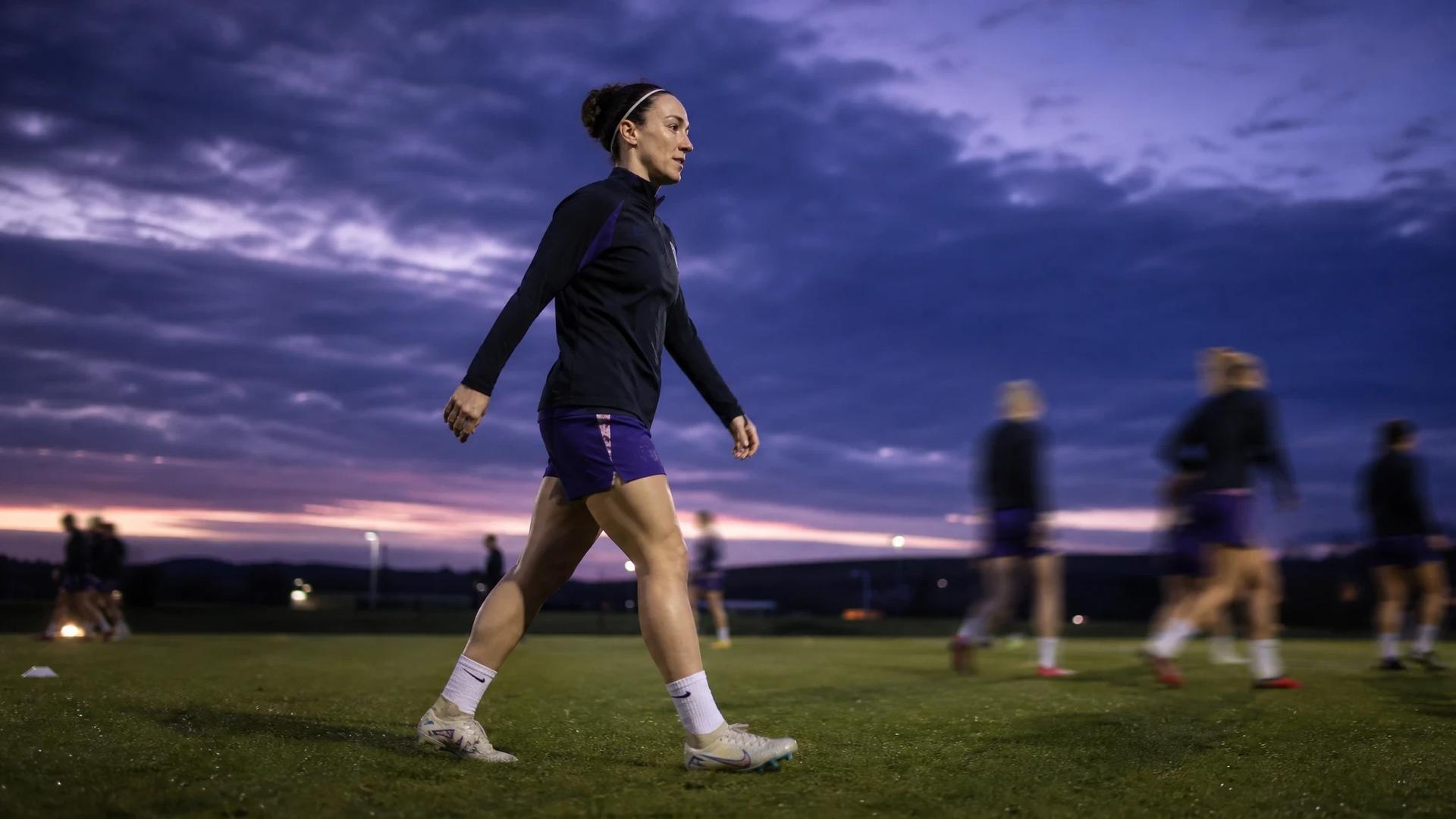 A determined Lucy Bronze walks across a training ground at dusk, with teammates in the background, symbolizing a pivotal moment in women's football.