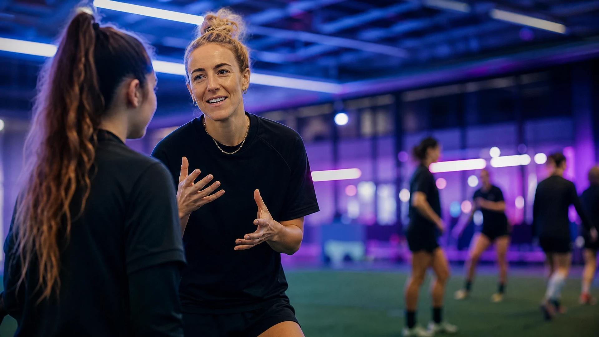 Carolin Simon engages in a tactical discussion with a younger teammate in a neon-lit training facility.