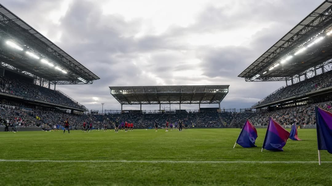 A wide view of a stadium with players warming up in the distance, under an overcast sky.