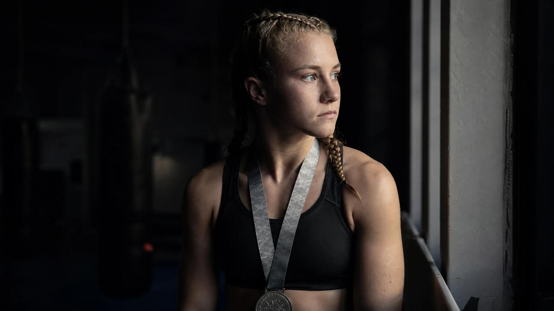 A young female boxer, Ruby White, stands in a gym holding a silver medal, reflecting on her boxing journey.
