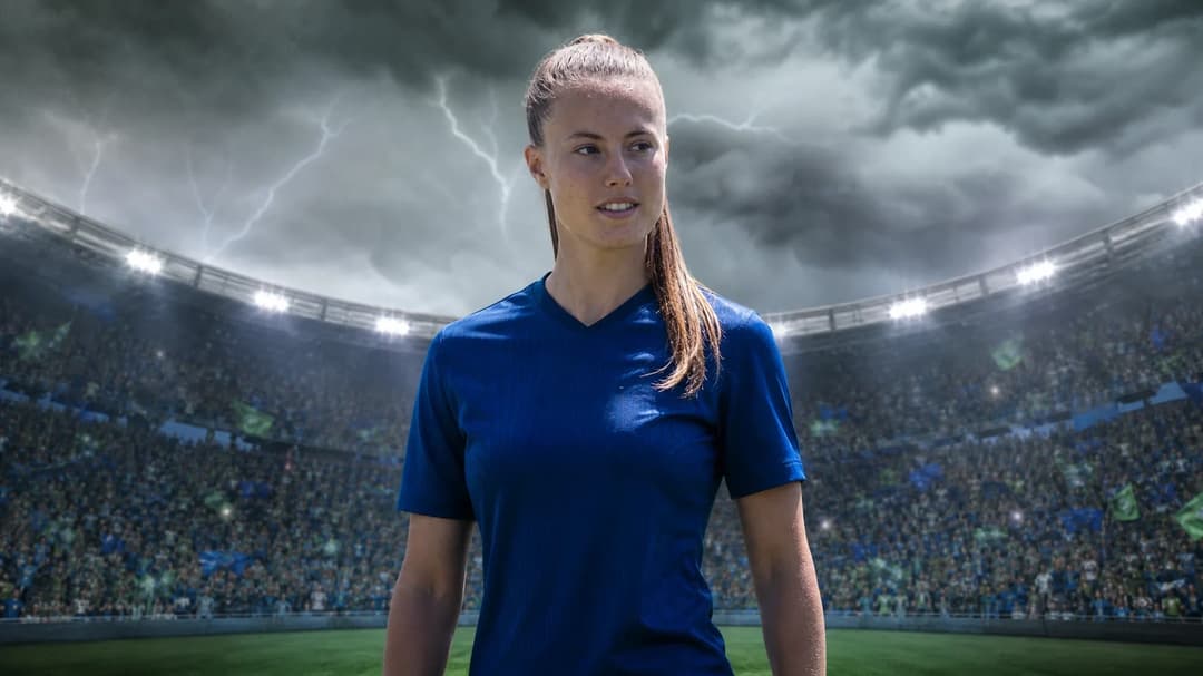 A young female footballer, Mara Alber, stands ready at the pitch edge under a stormy sky, with a crowd behind her.