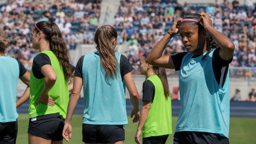A female football player adjusting her hairband in a sunny stadium with teammates in the background.