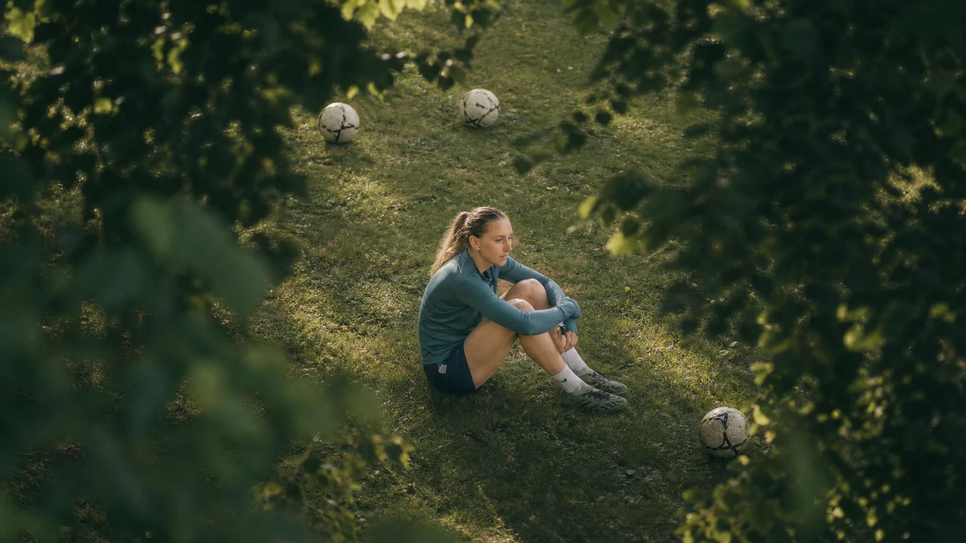 A bird's-eye view of a female football player sitting on the grass, reflecting in a serene training ground.