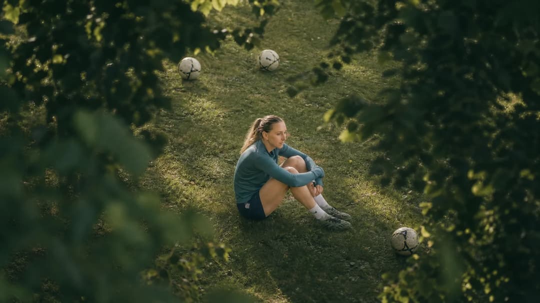 A bird's-eye view of a female football player sitting on the grass, reflecting in a serene training ground.