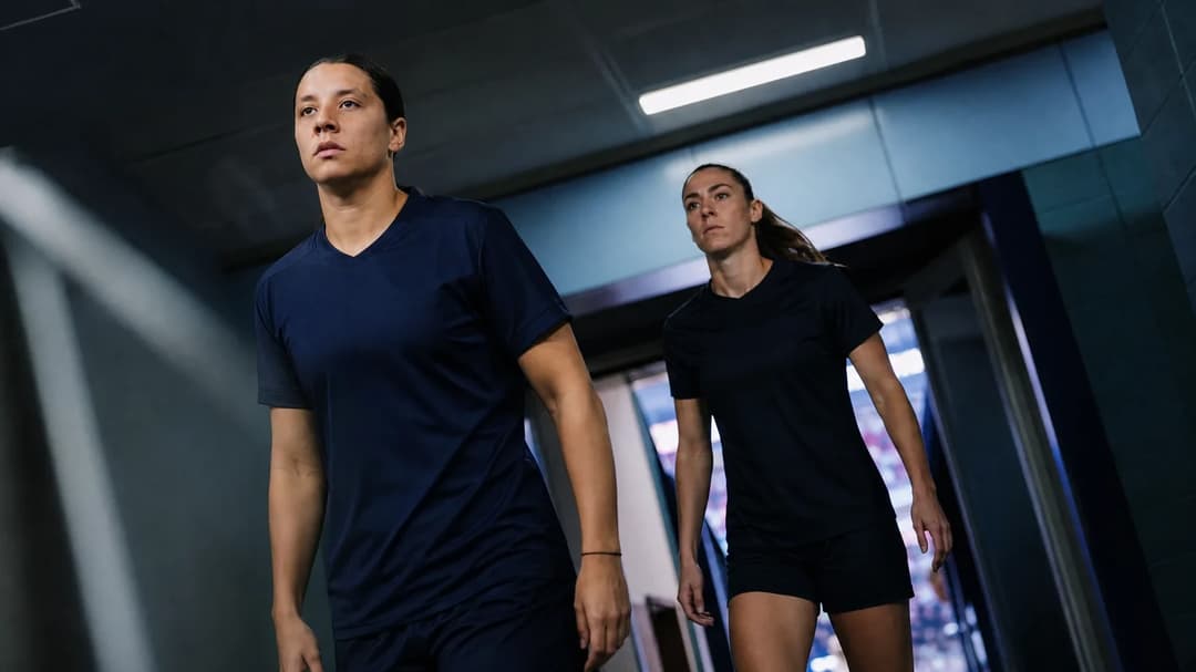 Two female football players transition from the locker room to the pitch, illuminated by cold fluorescent light.