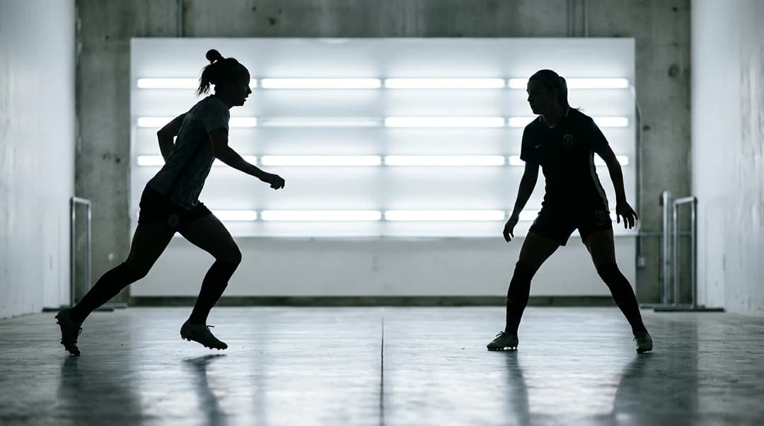 Two female football players in training interact against a bright fluorescent backdrop, captured as silhouettes.