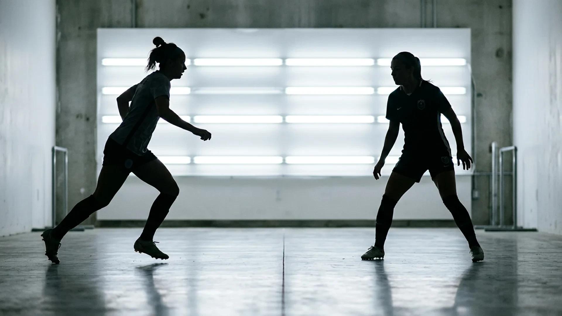 Two female football players in training interact against a bright fluorescent backdrop, captured as silhouettes.