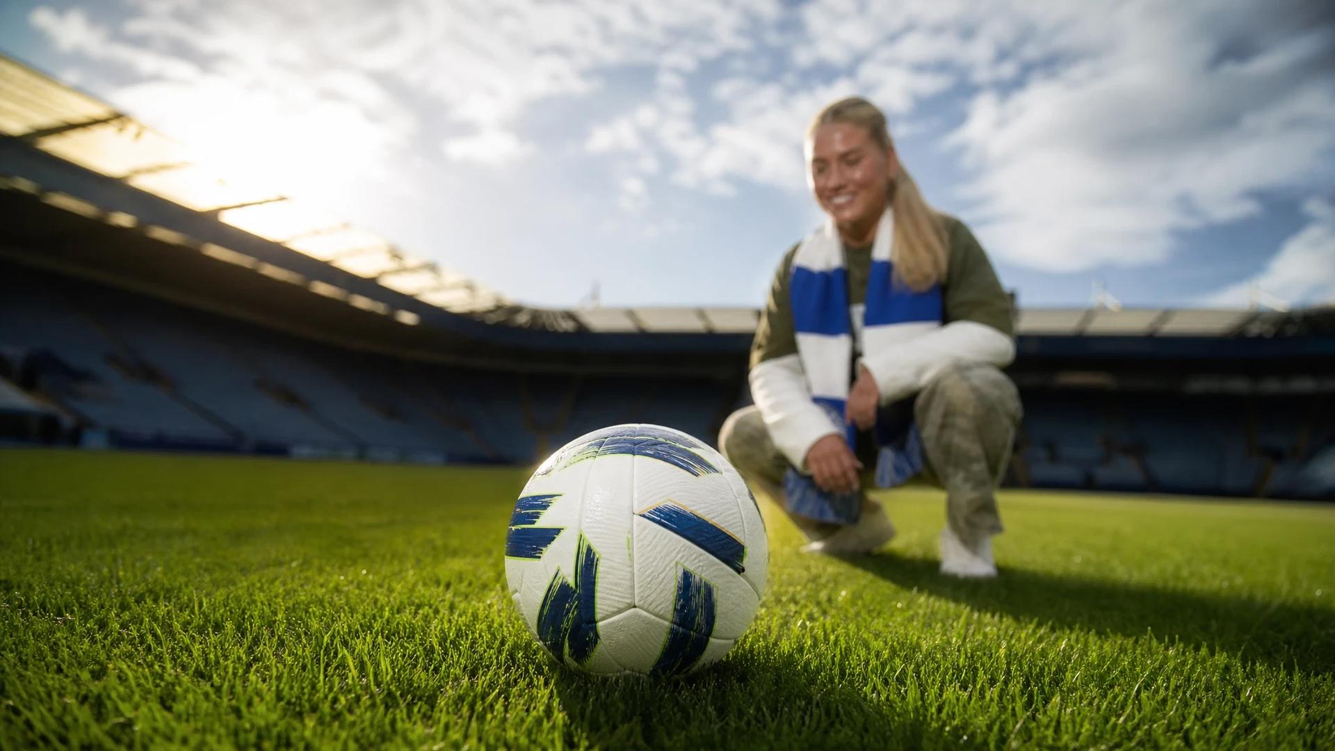 A close-up of a football on grass with a backdrop of a blurred player preparing for a match.