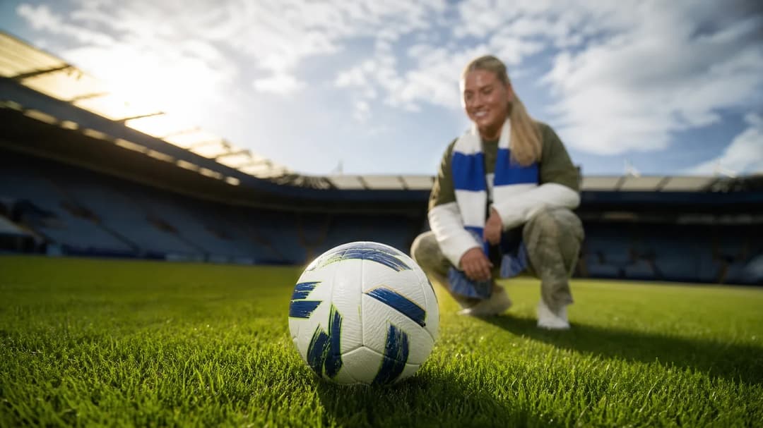 A close-up of a football on grass with a backdrop of a blurred player preparing for a match.
