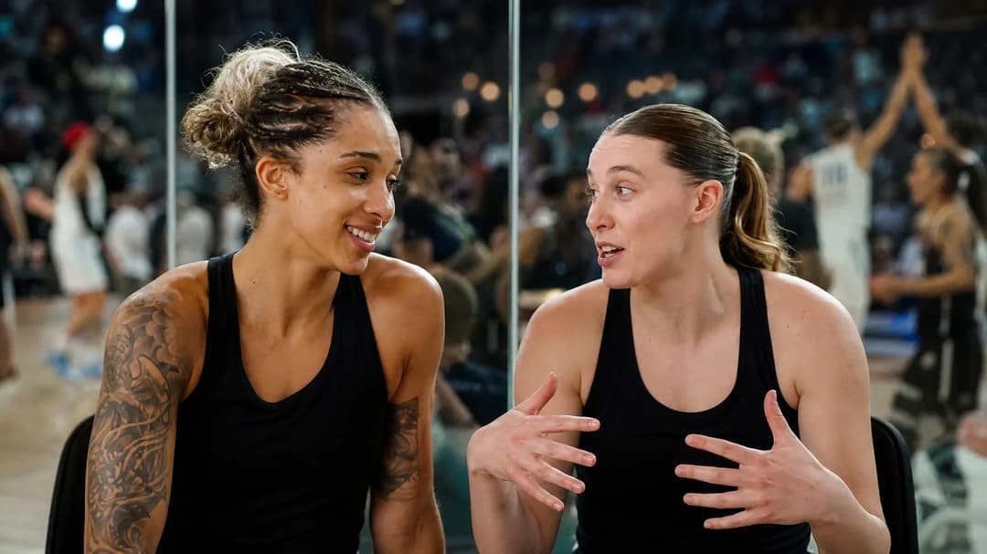 Gabby Williams and Veronica Burton share a moment of celebration in front of a glass wall reflecting the game.