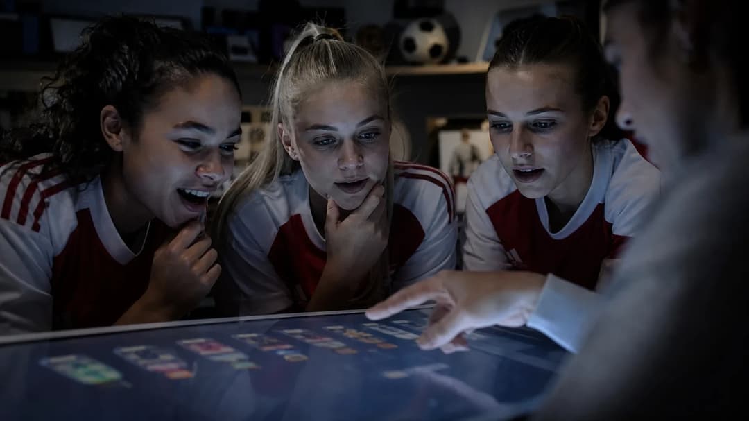 A focused group of young women discussing their football line-ups in front of a digital screen.
