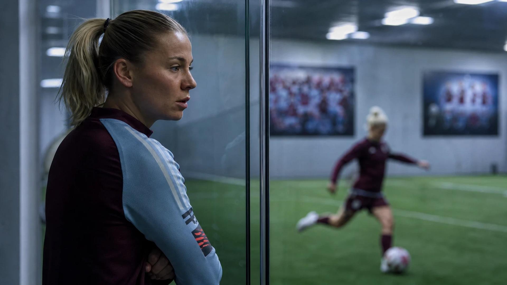 A young female football player in conversation, with another practicing free kicks in the background, captured through a glass partition.