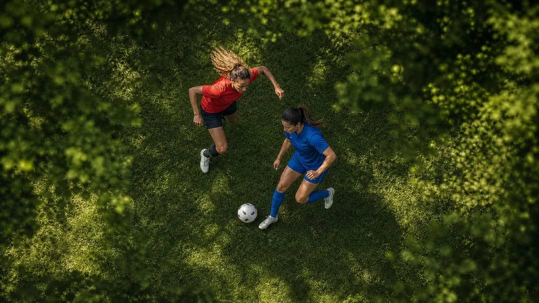 Aerial view of two female football players interacting on the pitch, surrounded by greenery.