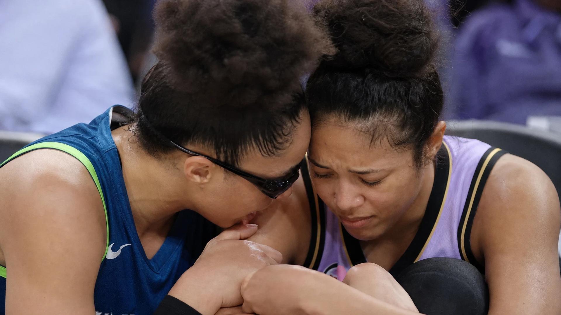 Close-up of two basketball players in a moment of concern and support, one sitting with an injured ankle.