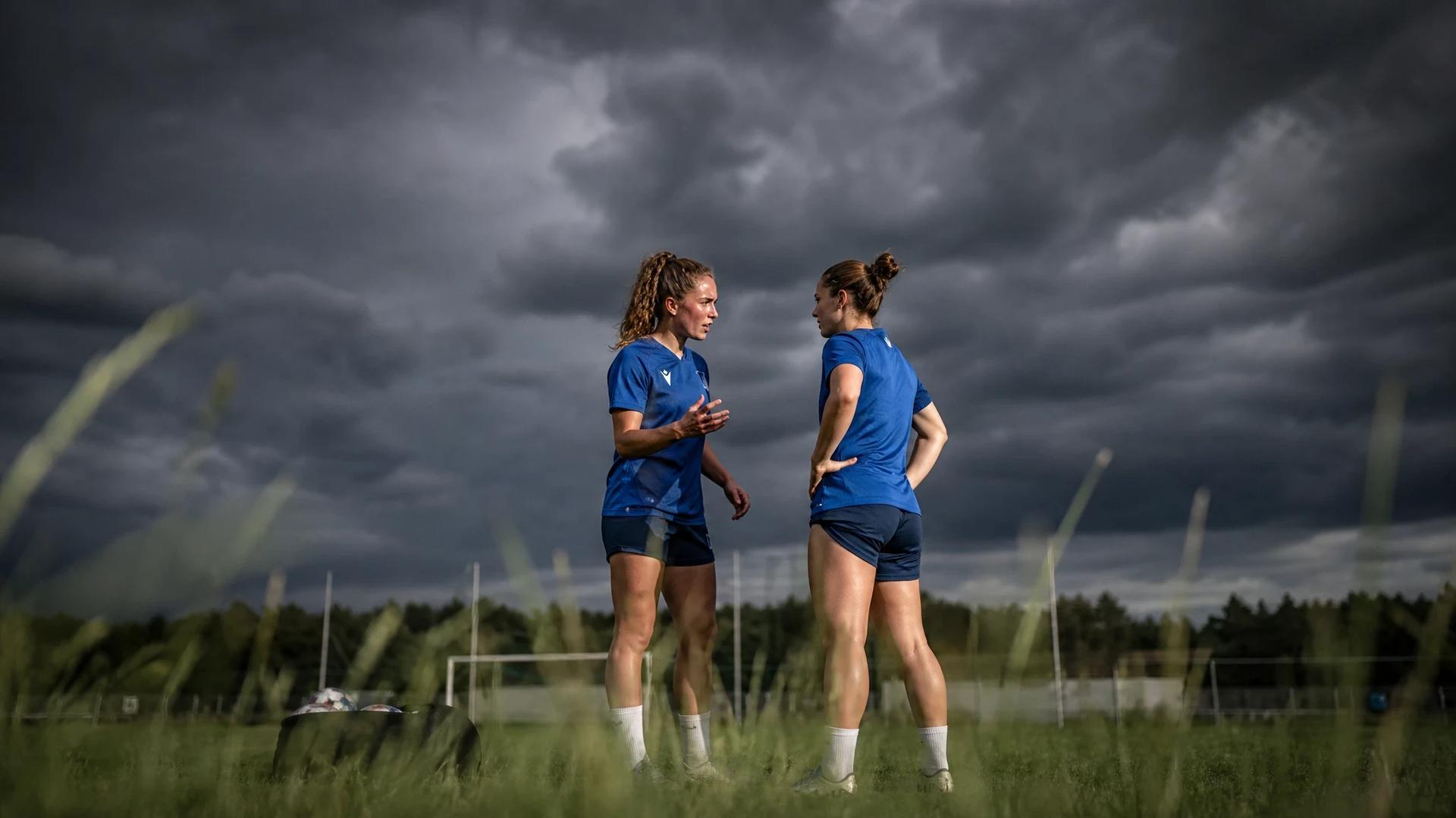 Two female football players from TSG Hoffenheim engaged in conversation on a stormy pitch.