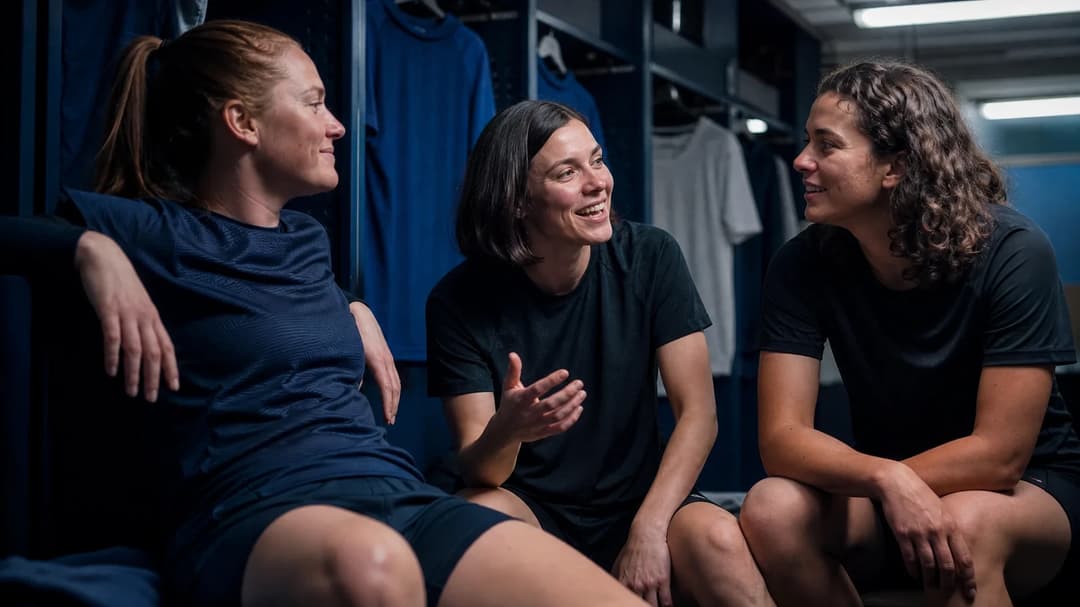Three female football players share a moment in a locker room, looking thoughtful and engaged.
