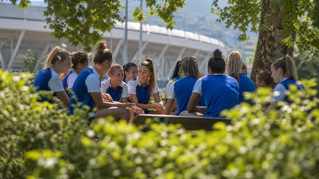 Juventus Women’s team sharing a candid moment before the match, seated on a bench surrounded by greenery.