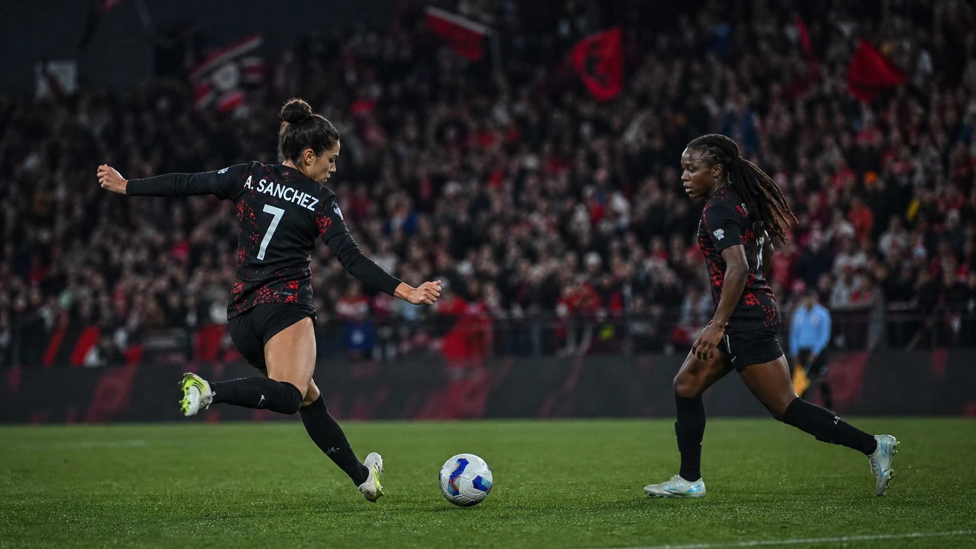 Two female football players from Portland Thorns in action during a match, with a cheering crowd in the background.