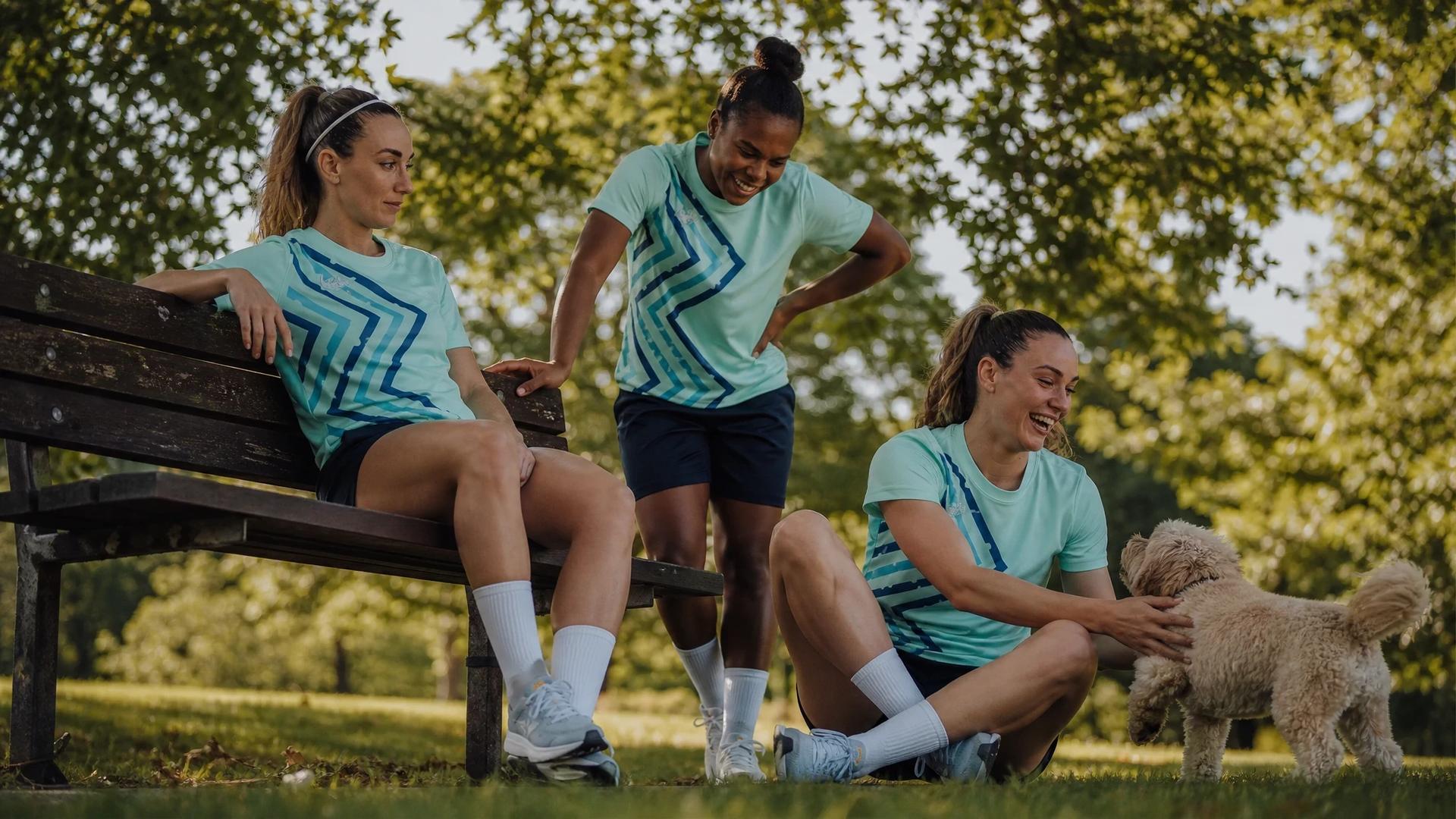 Three female football players enjoy a moment together in a park, with sunlight filtering through the leaves above.