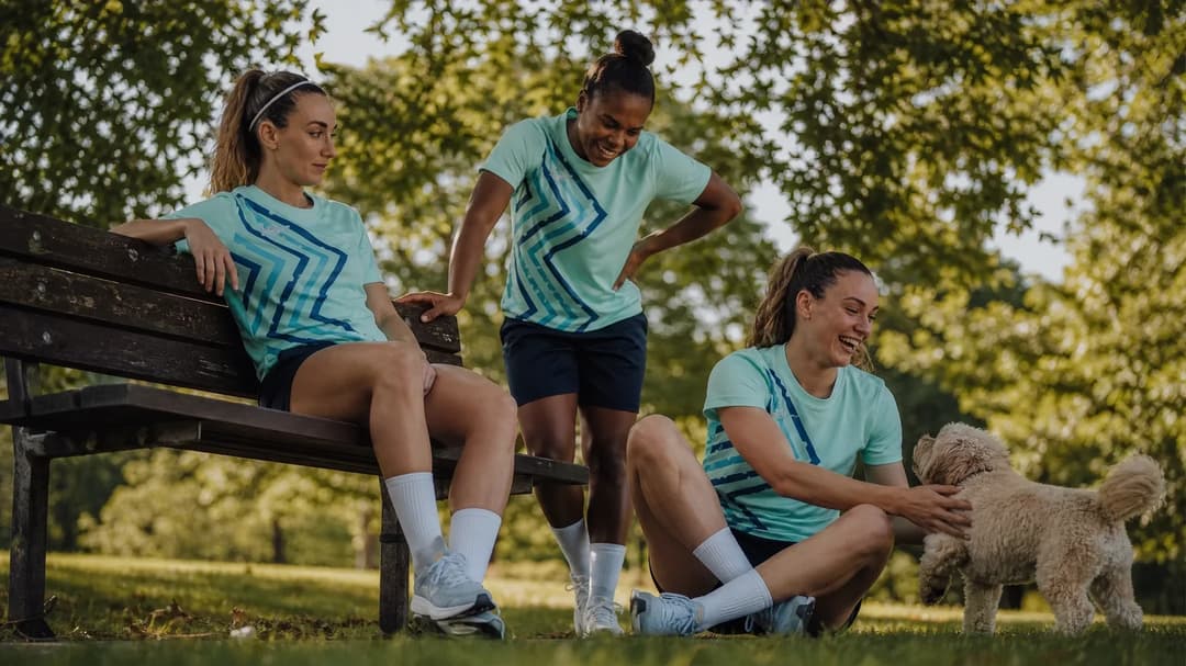 Three female football players enjoy a moment together in a park, with sunlight filtering through the leaves above.