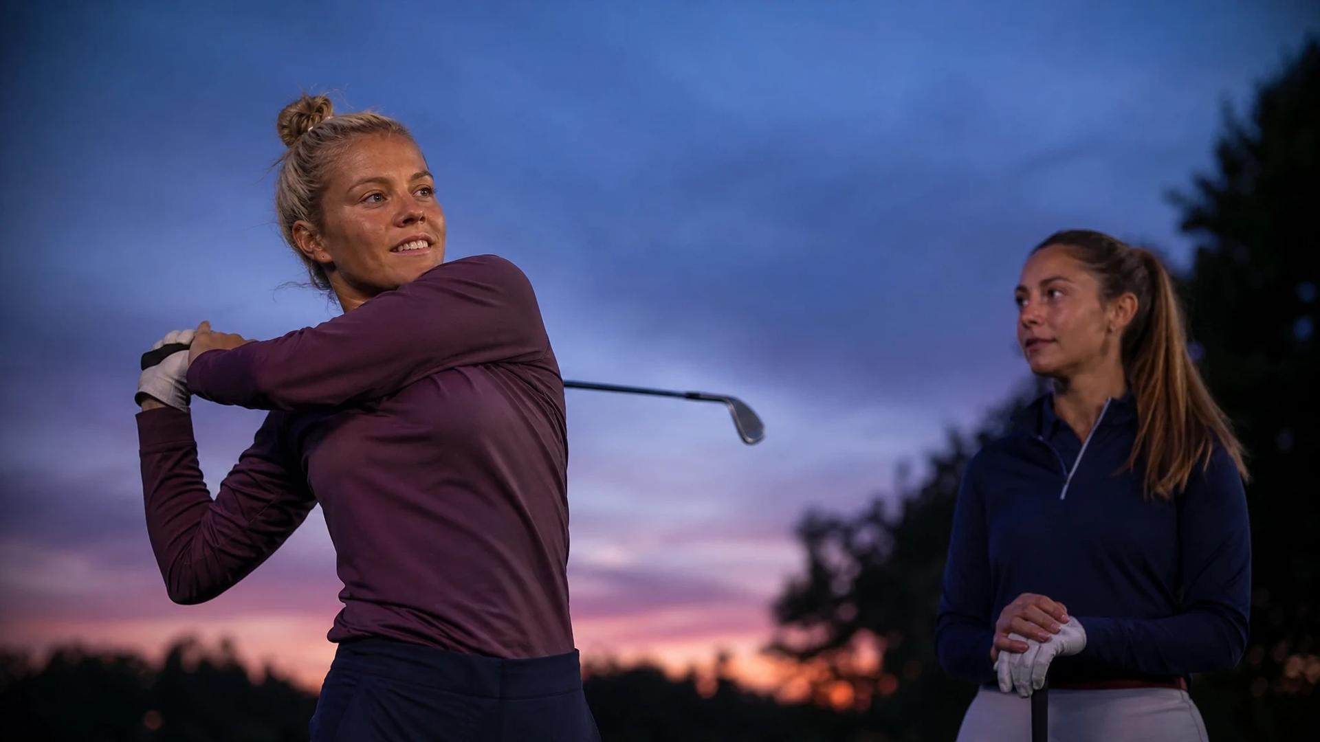 Rachel Daly swings her golf club with a teammate watching on a golf course at dusk.
