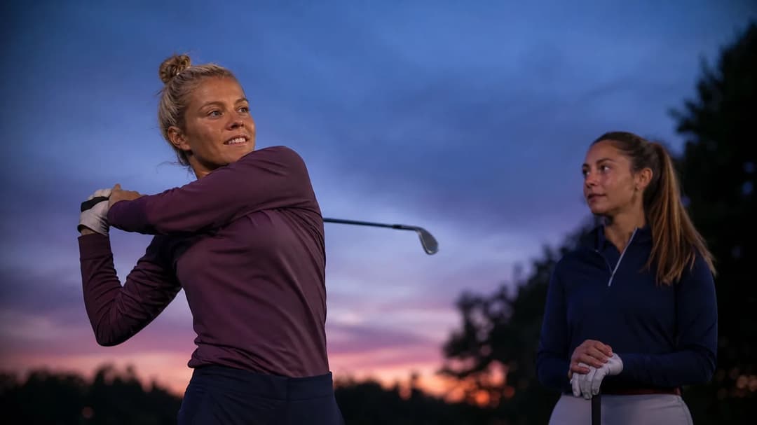Rachel Daly swings her golf club with a teammate watching on a golf course at dusk.