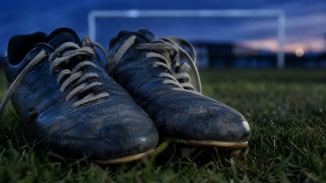 A close-up of worn football boots resting on grass, illuminated by dusk light.