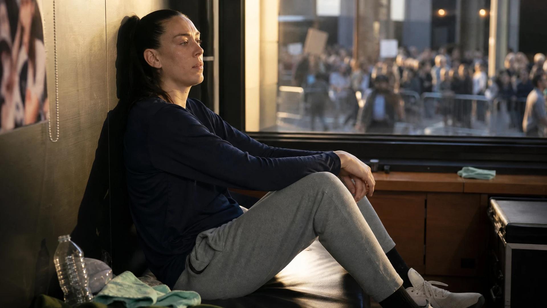 Breanna Stewart sitting thoughtfully in a dimly lit players' lounge, gazing out a window while dressed in sports attire.