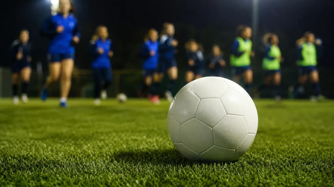 A pristine football sits on green grass, with blurred players training in the background.