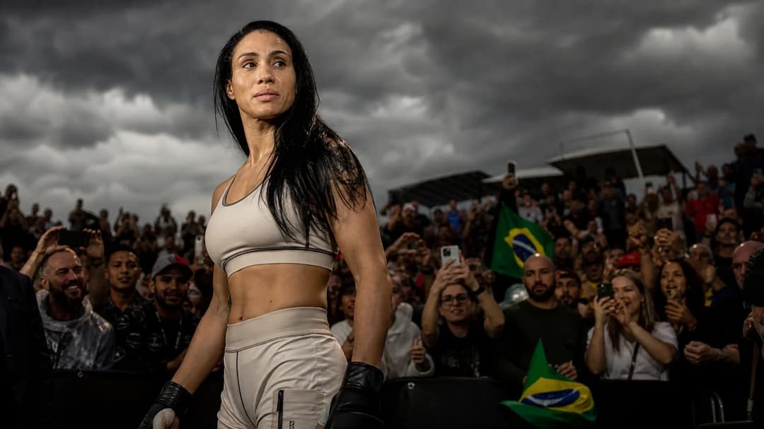 A female fighter, Taila Santos, stands confidently in the center of a bustling outdoor arena, amidst an excited crowd, under dramatic storm clouds.