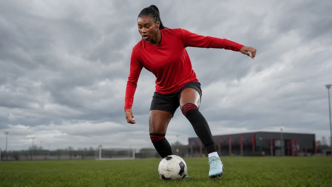 A determined Monica Jusu Bah poised on a training ground, with a close-up of her football cleats in action, against a blurred training facility backdrop.