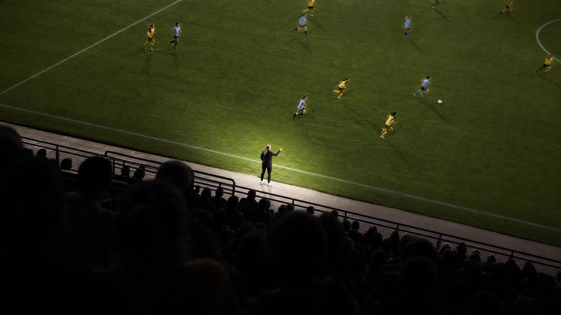 A bird's-eye view of a football match showing a female coach directing her team, capturing a dynamic moment of play.