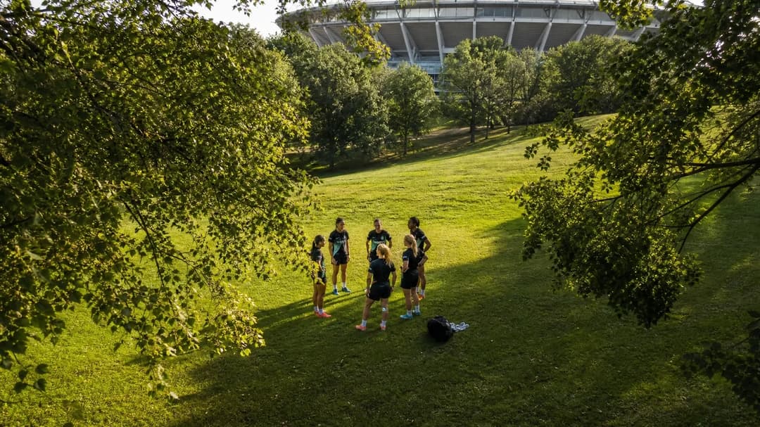 A top-down view of NJ/NY Gotham FC players huddled together on the grass under dappled sunlight.