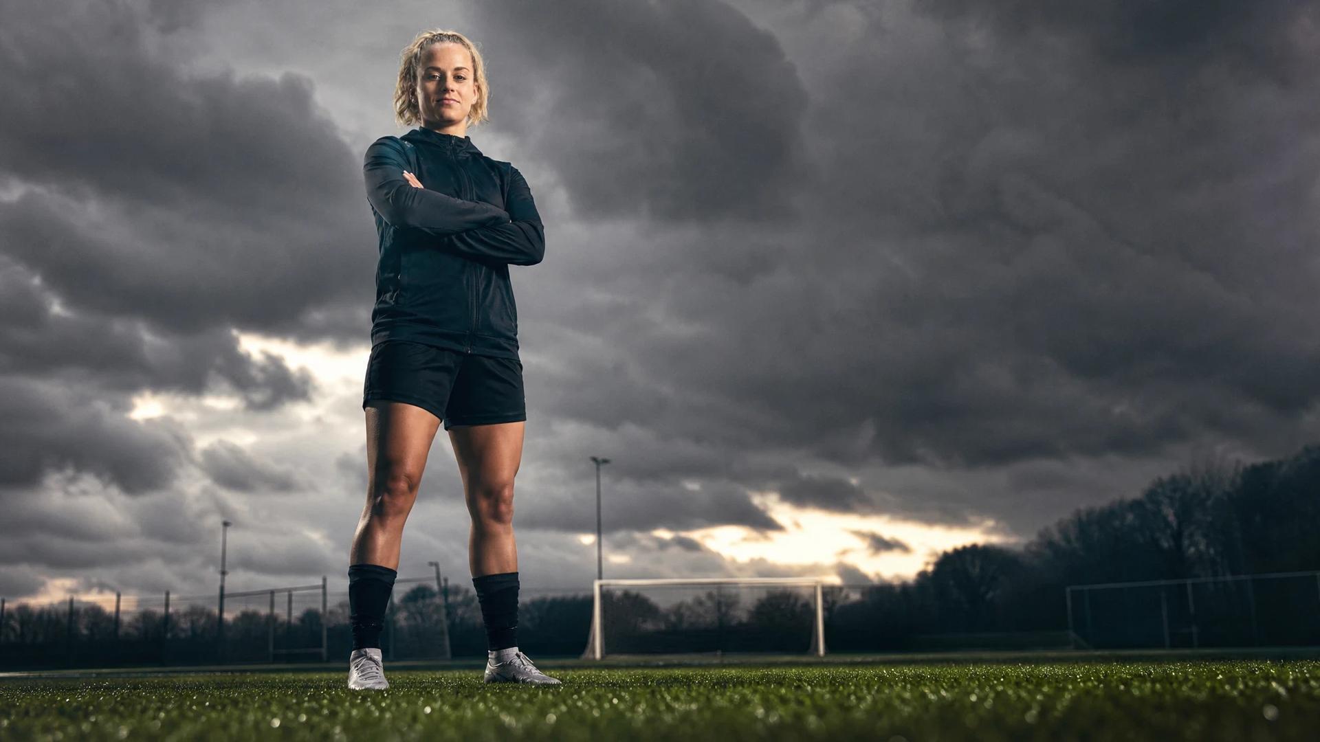 A female football player, Jenny Hipp, stands on a training pitch under a stormy sky.