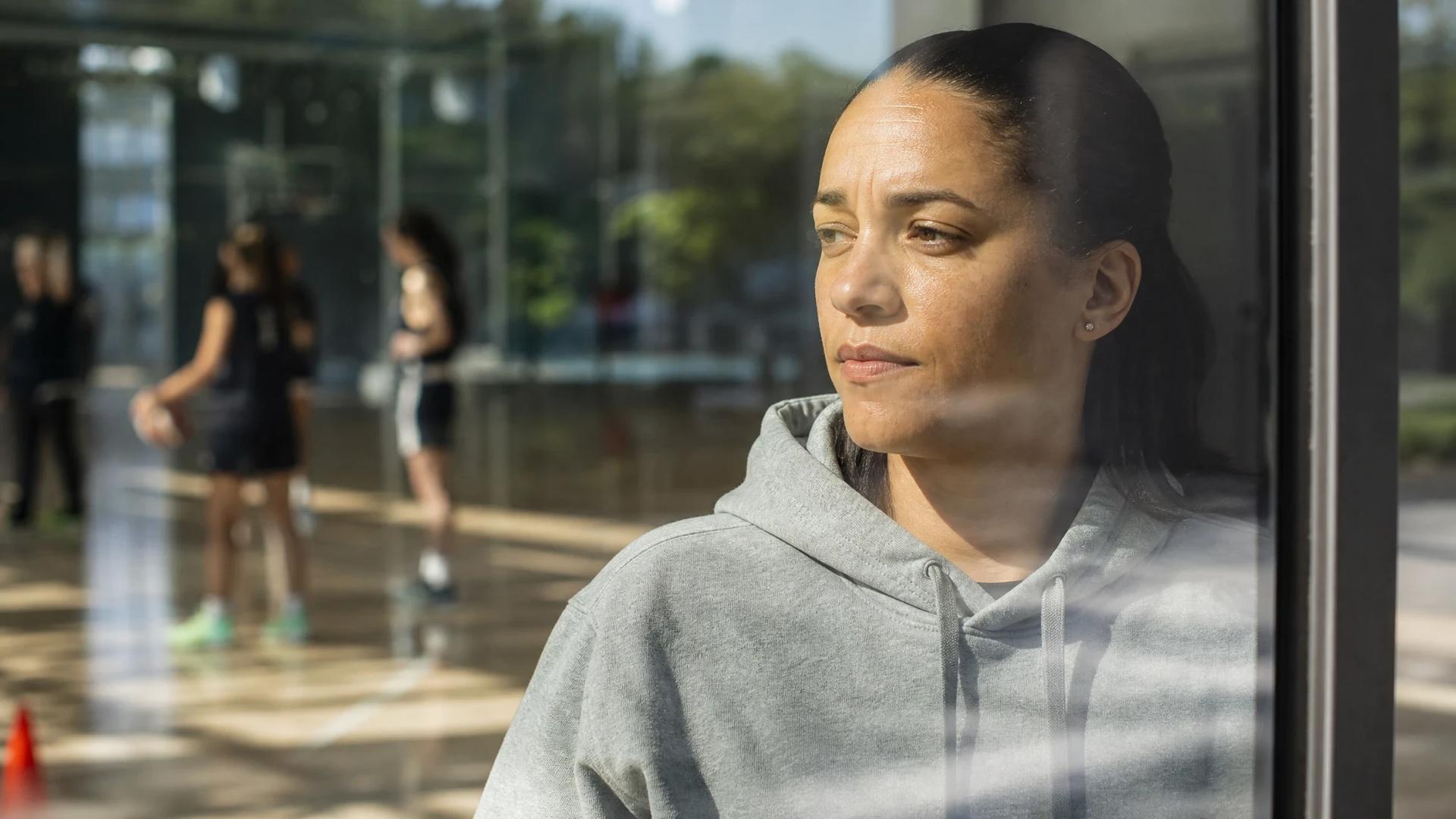 A contemplative basketball coach stands by a window, reflecting on her team's preparations.