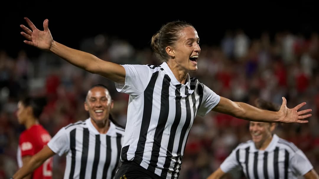 Woman in sports clothing celebrating a goal on a football pitch with cheering fans in the background.