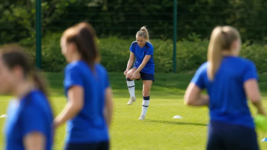 Grace Neville adjusting her shin guards on the training ground, with teammates blurred in the background.