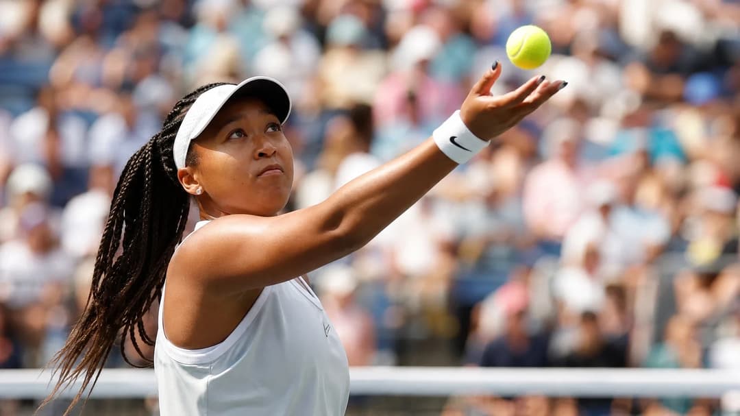 A tennis player, Naomi Osaka, is seen from an over-the-shoulder angle, preparing to serve on a vibrant court at the US Open.