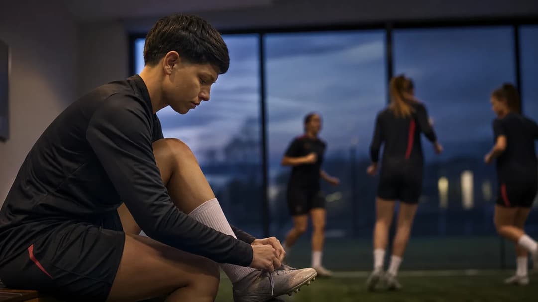 Élisa de Almeida tying her shoelaces in preparation for training, with teammates blurred in the background.