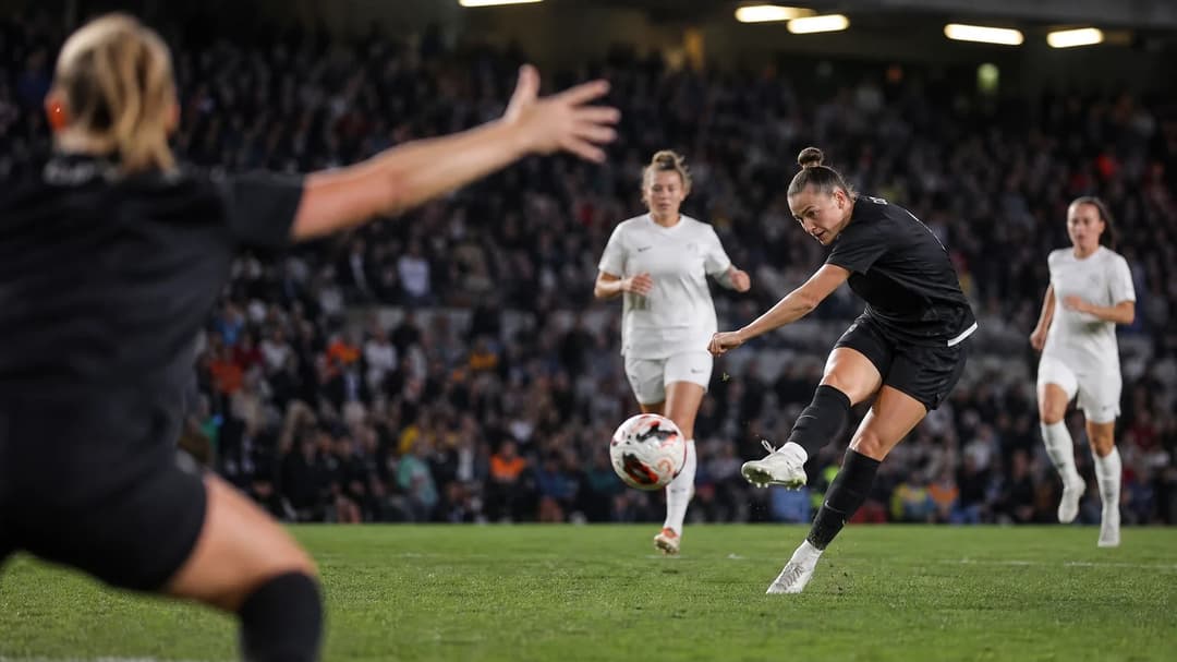 A female football player striking the ball during a match, with supporters in the background.